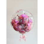 Clear balloon with pink balloons and "Happy Birthday" text, tied with a pink ribbon.