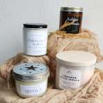 Four scented candles arranged on beige fabric with dried wheat accents.