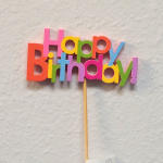Colorful "Happy Birthday!" sign on a stick against a plain wall