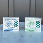 Two Honey Hill Farm bar soap boxes, Clean Rain and Rosemary Mint, on a light stone surface
