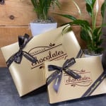 Two gold chocolate gift boxes tied with black ribbons beside potted plants
