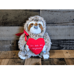 Brown sloth plush toy holding a red heart pillow with a message and red ribbon on a wooden surface