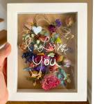 White framed shadow box with colorful dried flowers and 'I love you' written on the glass