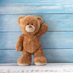 Brown plush teddy bear standing and waving in front of a blue wooden background