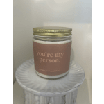 Glass jar candle with gold lid and label reading "you’re my person" on a white pedestal stand.