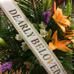 White memorial ribbon reading 'Dearly Beloved' draped over an arrangement with orange lilies.