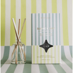 Reed diffuser in a clear glass bottle beside a striped product box