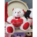 White teddy bear holding a red heart pillow with other plush bears nearby