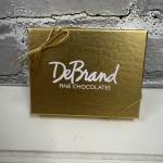 Gold DeBrand Fine Chocolates gift box with gold cord bow on a light surface