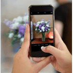 Hands holding a phone photographing a purple and white flower arrangement