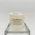 White scented candle labeled Peony White Tea in a clear glass jar on a glass stand
