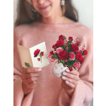 Woman in a pink sweater holding a rose-themed greeting card and a tiny bouquet in a patterned teacup.