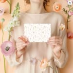 Person in a pale dress holding a floral greeting card surrounded by pastel flowers.