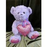 Lavender teddy bear holding a pink heart pillow with a ribbon