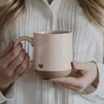 Two people holding a beige ceramic mug with a small heart cutout