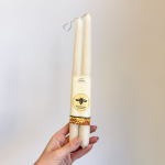 Hand holding two cream-colored taper candles in a paper sleeve