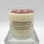 Glass jar candle labeled Cranberry Orange Zest with cream wax and red top layer on a clear stand