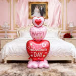 Heart-shaped Valentine's Day balloons stacked on a bed in a pink bedroom