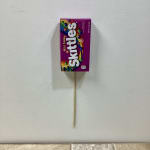 Box of wild berry Skittles candy attached to a wooden stick against a plain background