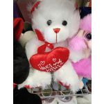 White teddy bear holding a red heart pillow with pink plush toys nearby