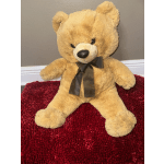 Tan teddy bear with a dark ribbon sitting on a red rug