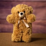 Fluffy light brown teddy bear plush with a satin bow on a brown surface.