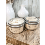 Two lidded tin candles labeled Black Currant + Jasmine on a wood block with white vases behind