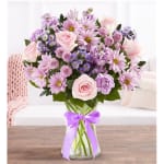 Pastel pink and purple mixed flower arrangement in a clear glass vase with a lavender ribbon.
