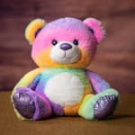 Rainbow plush teddy bear sitting on a brown surface