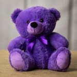 Seated purple teddy bear plush with matching satin bow on a neutral background