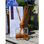 Glass reed diffuser with amber oil in front of a Rosy Rings box and blue flowers.