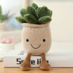 Smiling beige plush pot with green succulent-style top and dangling legs sitting on a book