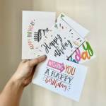 Hand holding four birthday greeting cards with colorful text and confetti