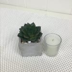 Small succulent in a square concrete pot with pebbles beside a white votive candle.