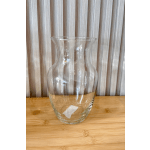Clear hourglass-shaped glass vase on a light wood surface against a striped wall.