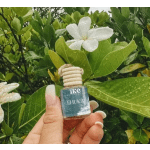 Hand holding a small glass car diffuser bottle in front of white flowers and green leaves