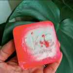 Hand holding a square pink-orange soap bar in front of large green leaves