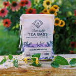 A tea bag pouch sits on a table with small flowers and leaves in front of a blurred flower field.