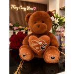 Large brown teddy bear holding a heart pillow in a flower shop