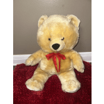 Large beige teddy bear with a red ribbon sitting on a red rug