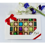 Box of assorted chocolates with red ribbon beside white and purple flowers