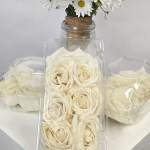 Cream roses in clear plastic boxes with a small jar vase behind them