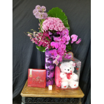 Pink roses and orchids in a purple vase with a teddy bear and chocolates