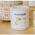 White scented candle in a glass jar labeled homebody on a wooden surface