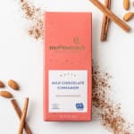 Coral box of nutty milk chocolate cinnamon bar with cinnamon sticks, almonds, and spice scattered around