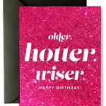 Pink glitter birthday card with white text saying older, hotter, wiser and a black envelope behind it