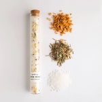 Glass tube of bath soak with three ingredient piles beside it