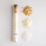Glass tube of bath soak with oats and salt beside loose piles of ingredients