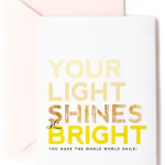 White greeting card with yellow and gold text reading 'Your light shines bright' and a pale pink envelope