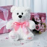 White teddy bear with a pink ribbon beside pink gift boxes and flowers